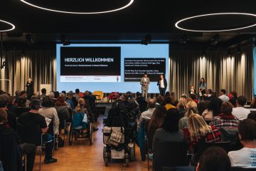 Blick auf ein breites Podium, zwei Männer stehen darauf, dahinter eine Powerpoint Präsentation und eingeblendete Schriftdolmetschung