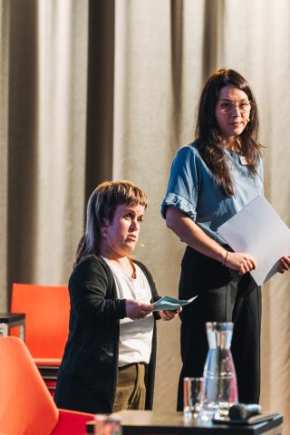 zwei Frauen mit Headset und Moderationskarten am Podium