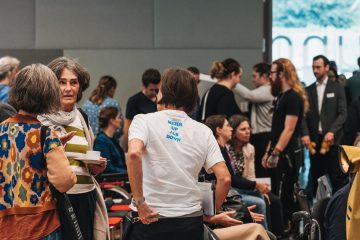 Eine Gruppe von Menschen in einem Veranstaltungsraum. Eine Person mit dem Rücken zur Kamera, auf dem T-Shirt steht "Mehr Up als Down"